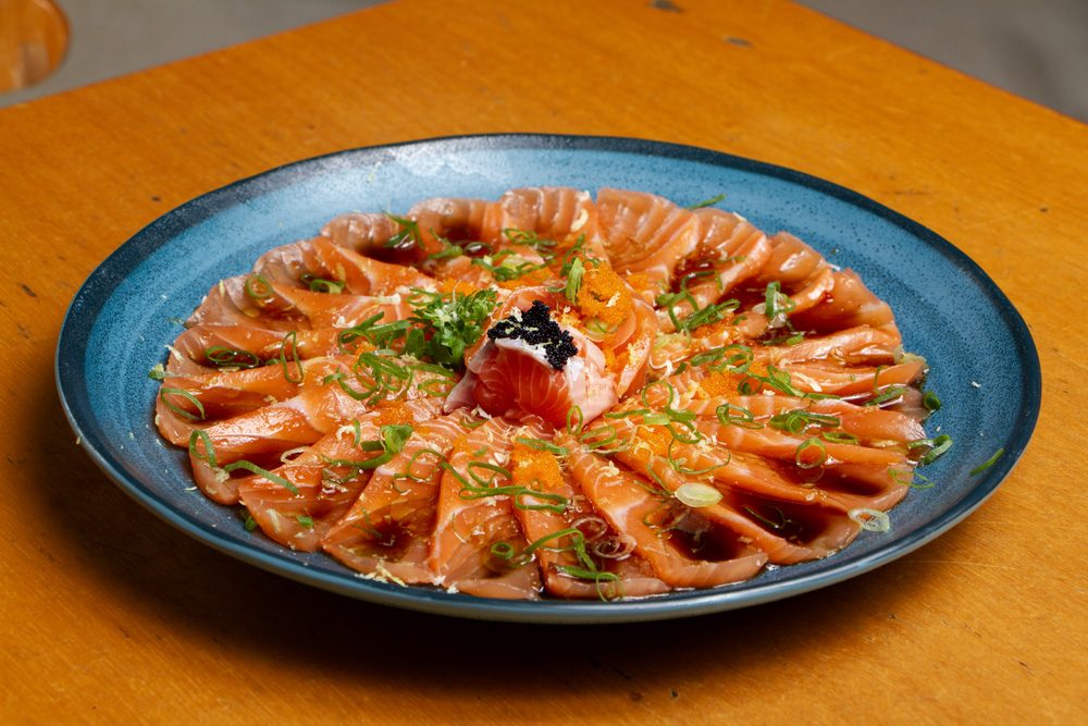 Carpaccio de salmão: lâminas finas de salmão cru dispostas em leque num prato azul, regadas com molho ponzu